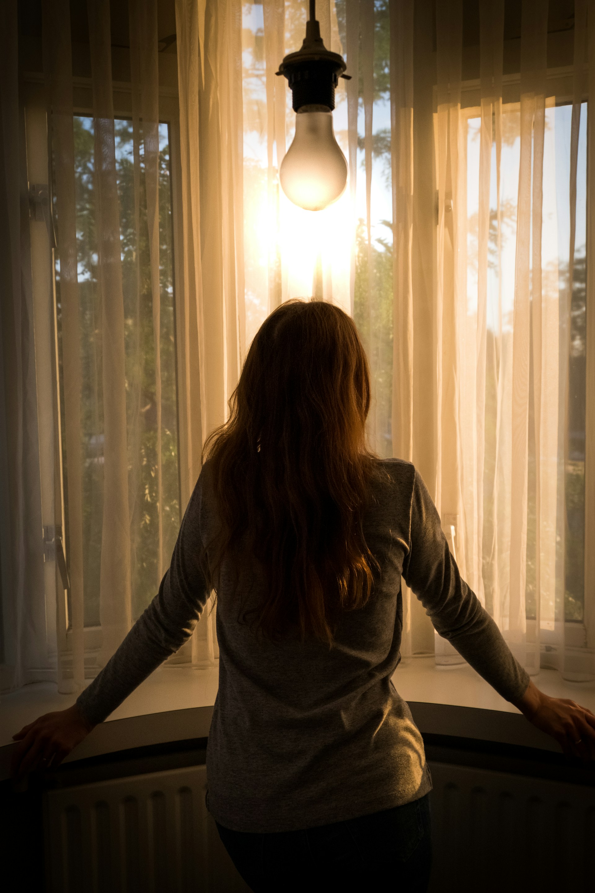 A young person standing alone by a window, backlit in soft natural light — a contemplative silhouette.